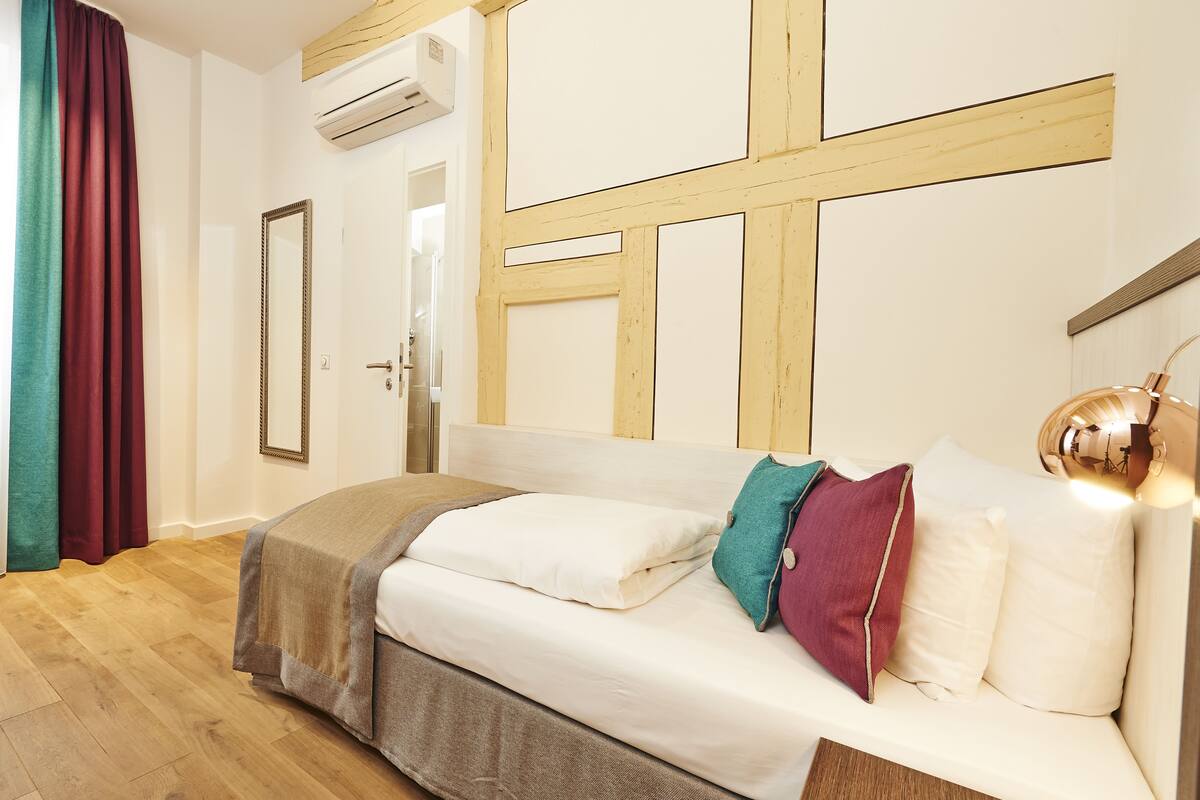 A hotel room with a wooden floor, a bed with white bedding and colorful pillows, a wooden headboard, and a large mirror.