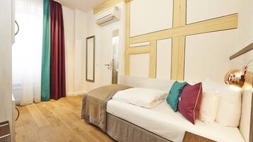 Habitación de hotel con piso de madera, cama con ropa blanca y almohadas coloridas, cabecera de madera y un espejo grande.