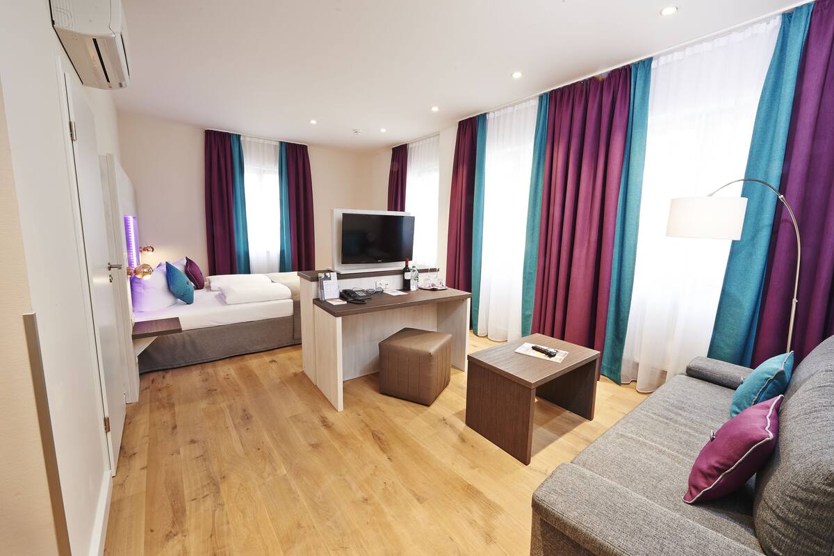 A modern hotel room with a sofa, a desk with a flat-screen TV, a small table, and a large window with curtains.
