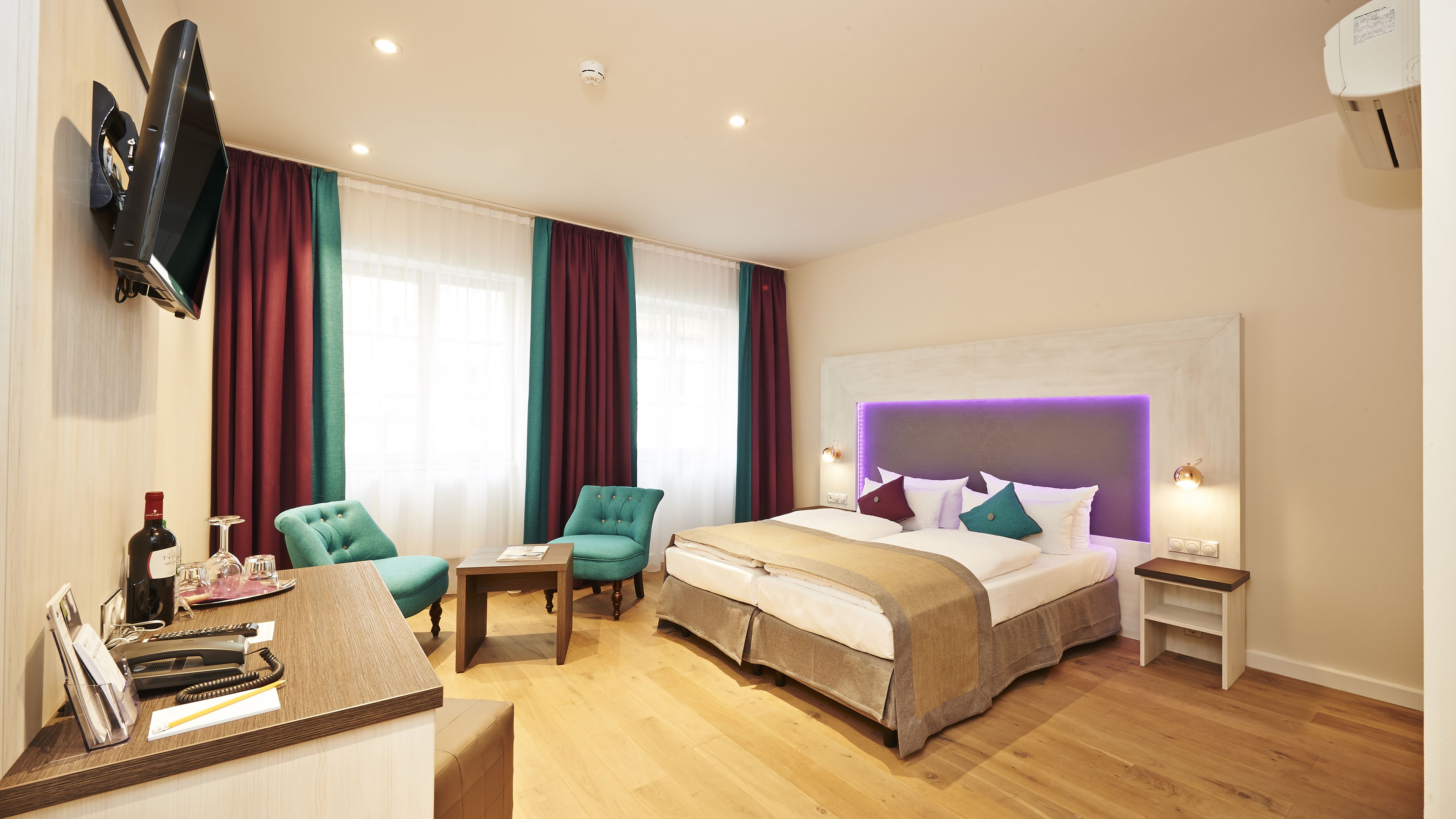 A hotel room with a bed, two armchairs, a desk with a telephone and wine bottle, and a TV mounted on the wall.