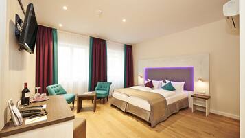 A hotel room with a bed, two armchairs, a desk with a telephone and wine bottle, and a TV mounted on the wall.