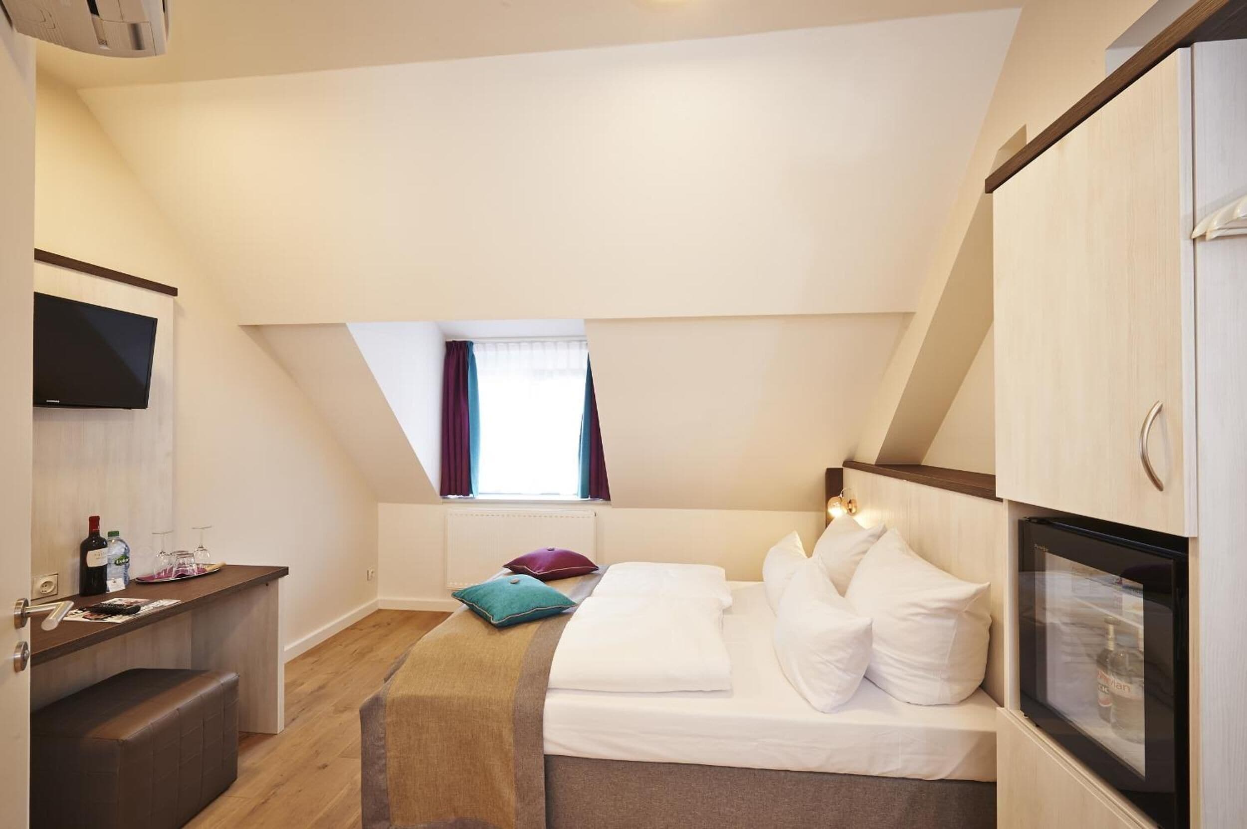 A compact hotel room with a bed, a small desk, a television, and a window with curtains.