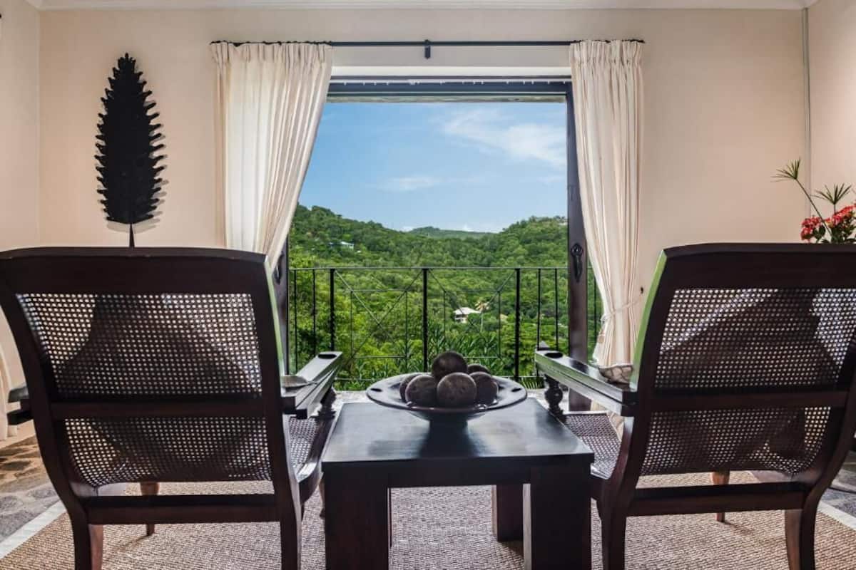 A balcony with two wicker chairs, a table with a plate of fruit, and a view of green hills.