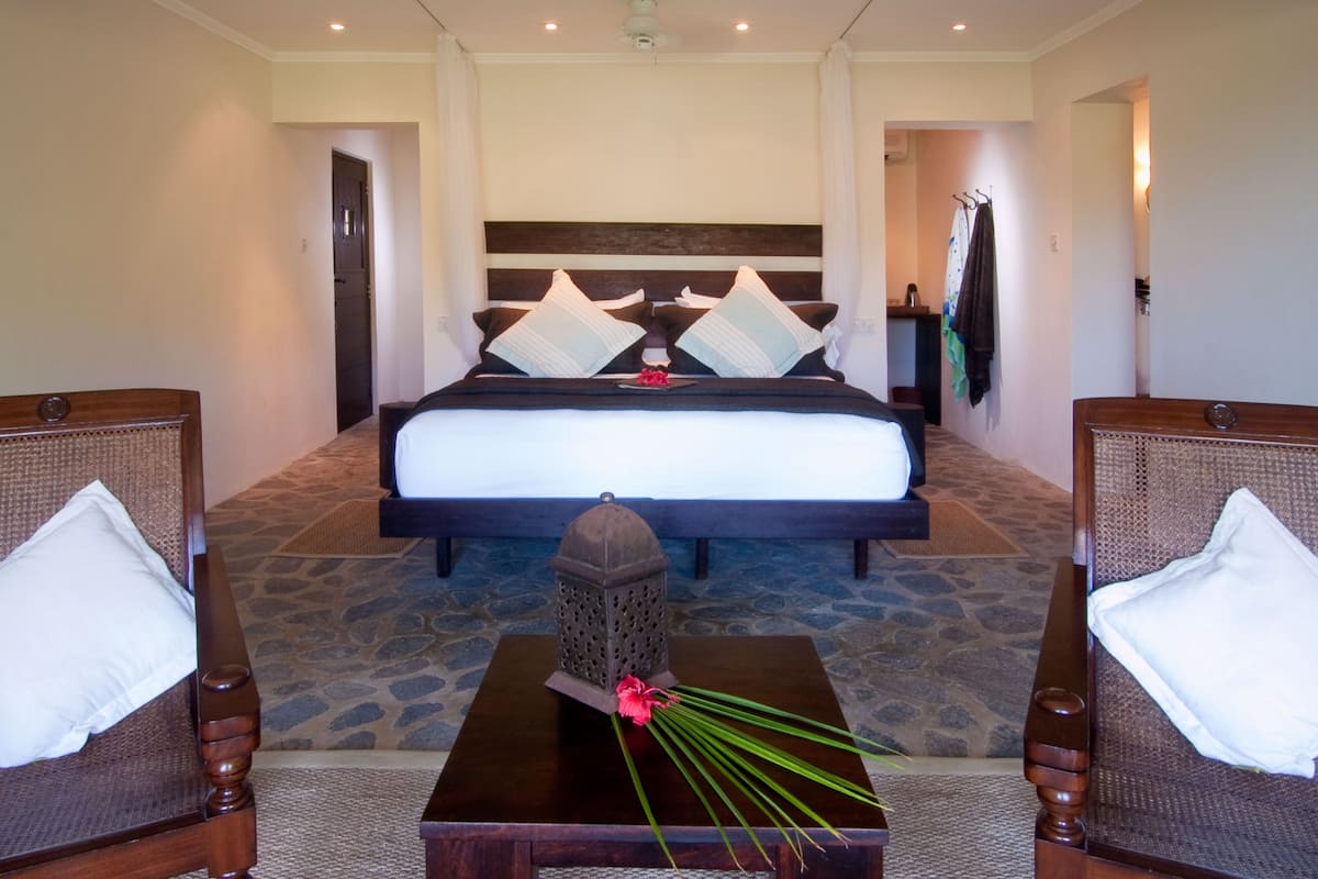 A hotel room with a large bed, two armchairs, a small table with a vase of flowers, and a stone-patterned floor.