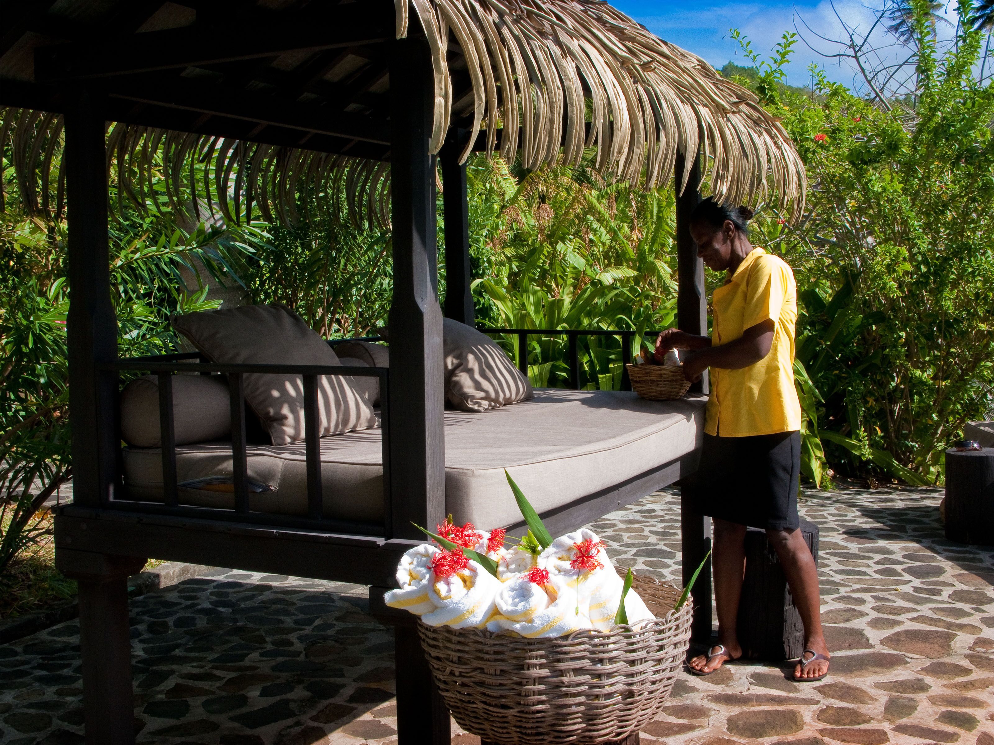 A thatched-roof outdoor seating area with a cushioned bench, a wicker basket, and a basket of flowers.