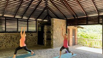 Two people practicing yoga on mats in a covered area with stone walls and a wooden ceiling.