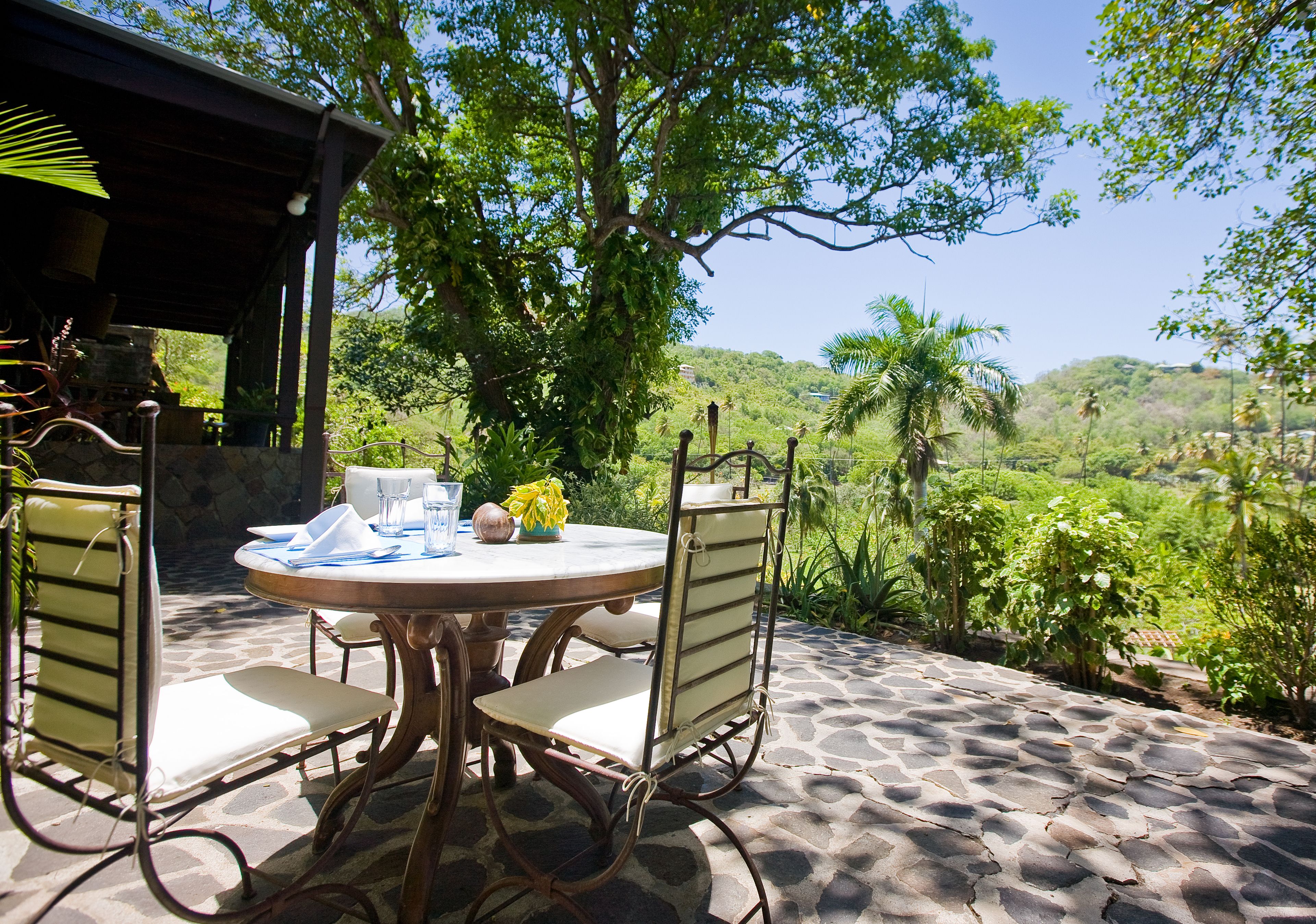 Un área de comedor al aire libre con una mesa redonda y sillas, rodeada de abundante vegetación y vistas a un paisaje montañoso.
