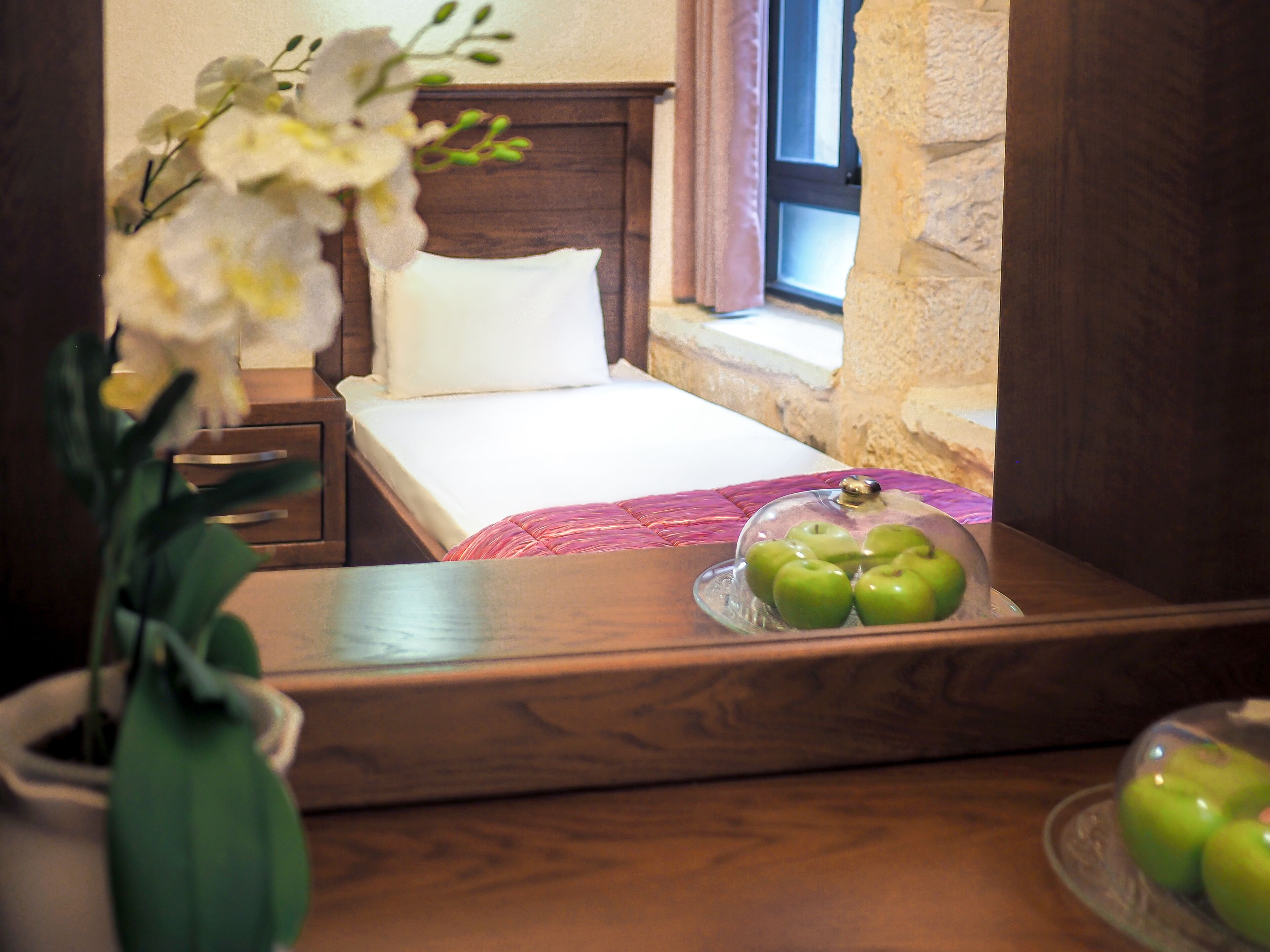 A neatly made bed with a white pillow and a pink blanket, a wooden nightstand with a vase of flowers, and a glass bowl of green apples on the dresser.
