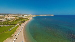 An aerial view of a coastal resort with a long sandy beach, rows of beach umbrellas, and a clear blue sea.