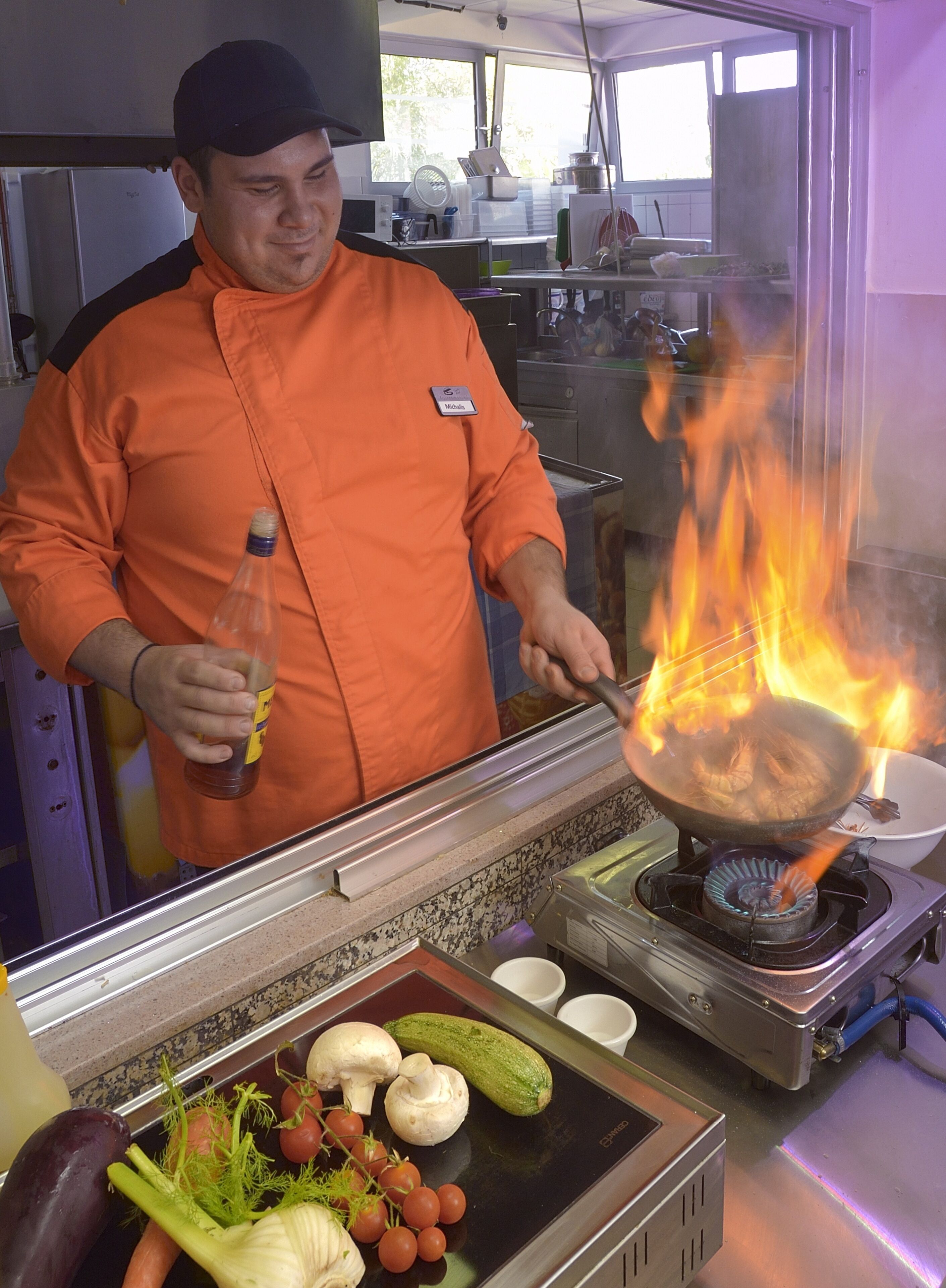 A chef in an orange uniform cooking with a wok over an open flame, surrounded by vegetables and cooking utensils.