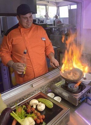 A chef in an orange uniform cooking with a wok over an open flame, surrounded by vegetables and cooking utensils.