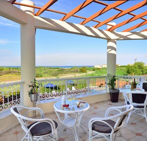 A covered outdoor area with white wicker chairs and a table, overlooking a scenic landscape with greenery and a distant view of the sea.