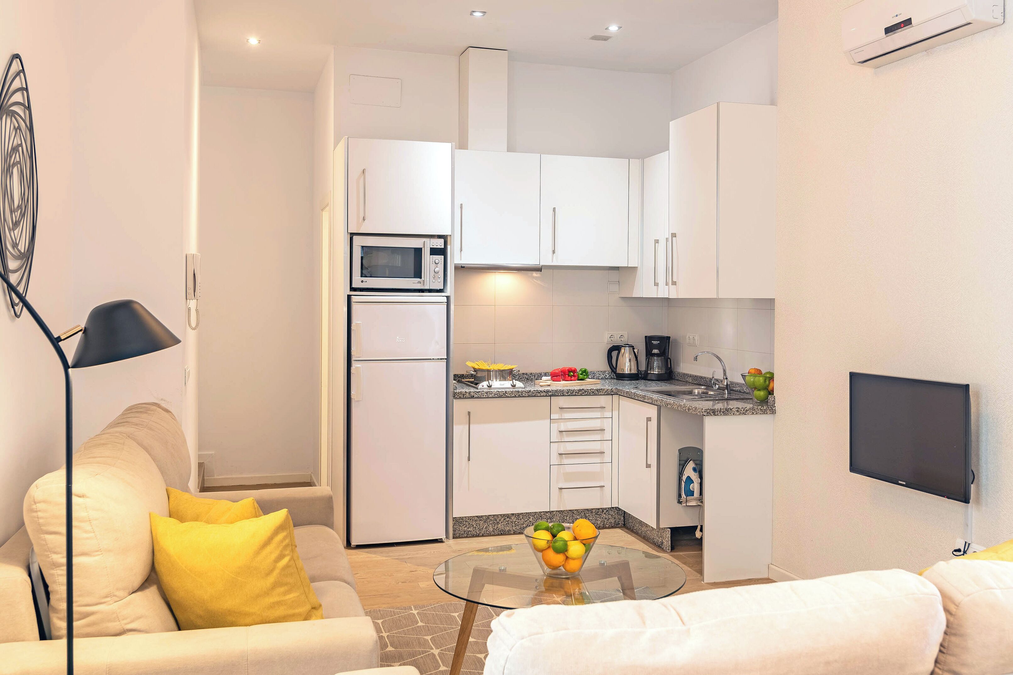 A modern kitchen with white cabinets, a microwave, and a coffee maker. There is a glass table with a fruit bowl in the center of the room.