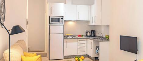 A modern kitchen with white cabinets, a microwave, and a coffee maker. There is a glass table with a fruit bowl in the center of the room.