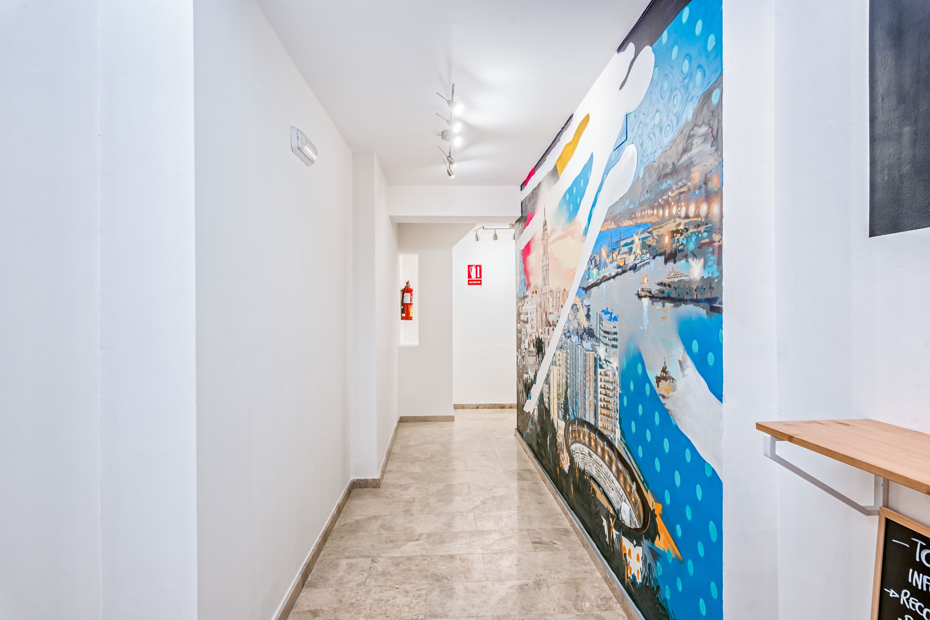A hallway with a cityscape mural on the right wall, a fire extinguisher on the left, and a wooden shelf on the right.