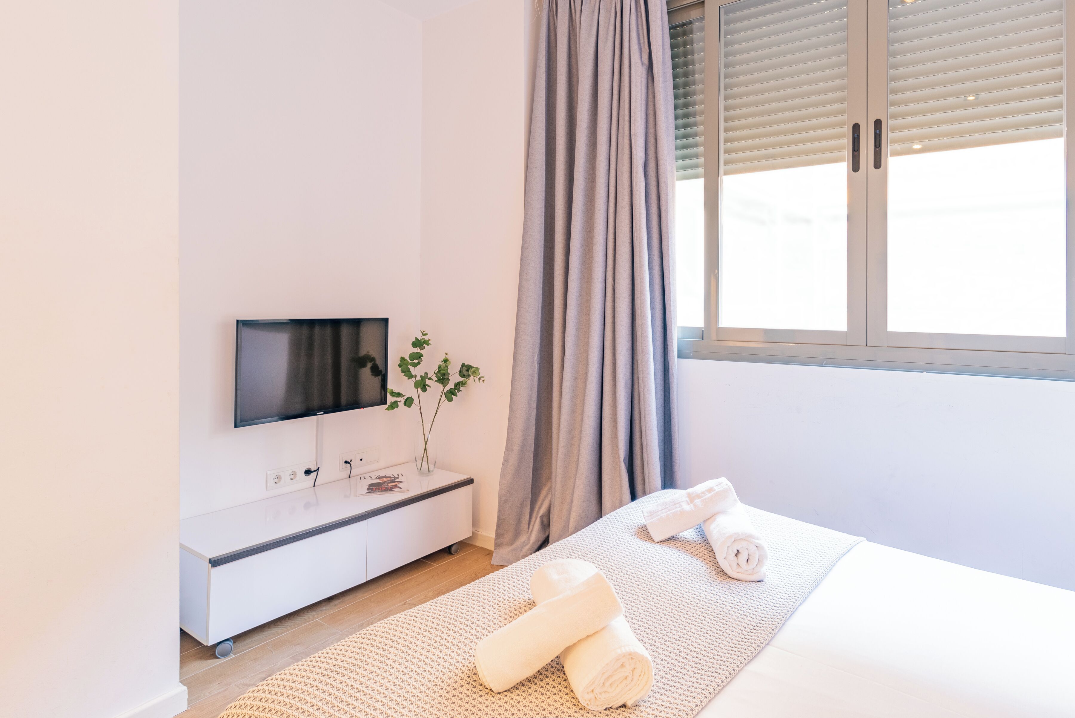 Een moderne hotelkamer met een flatscreen televisie aan de muur, een nachtkastje met een plant, een groot raam met gordijnen, en netjes opgevouwen handdoeken op het bed.