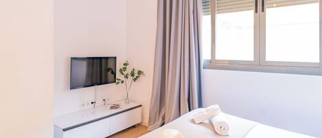 Een moderne hotelkamer met een flatscreen televisie aan de muur, een nachtkastje met een plant, een groot raam met gordijnen, en netjes opgevouwen handdoeken op het bed.