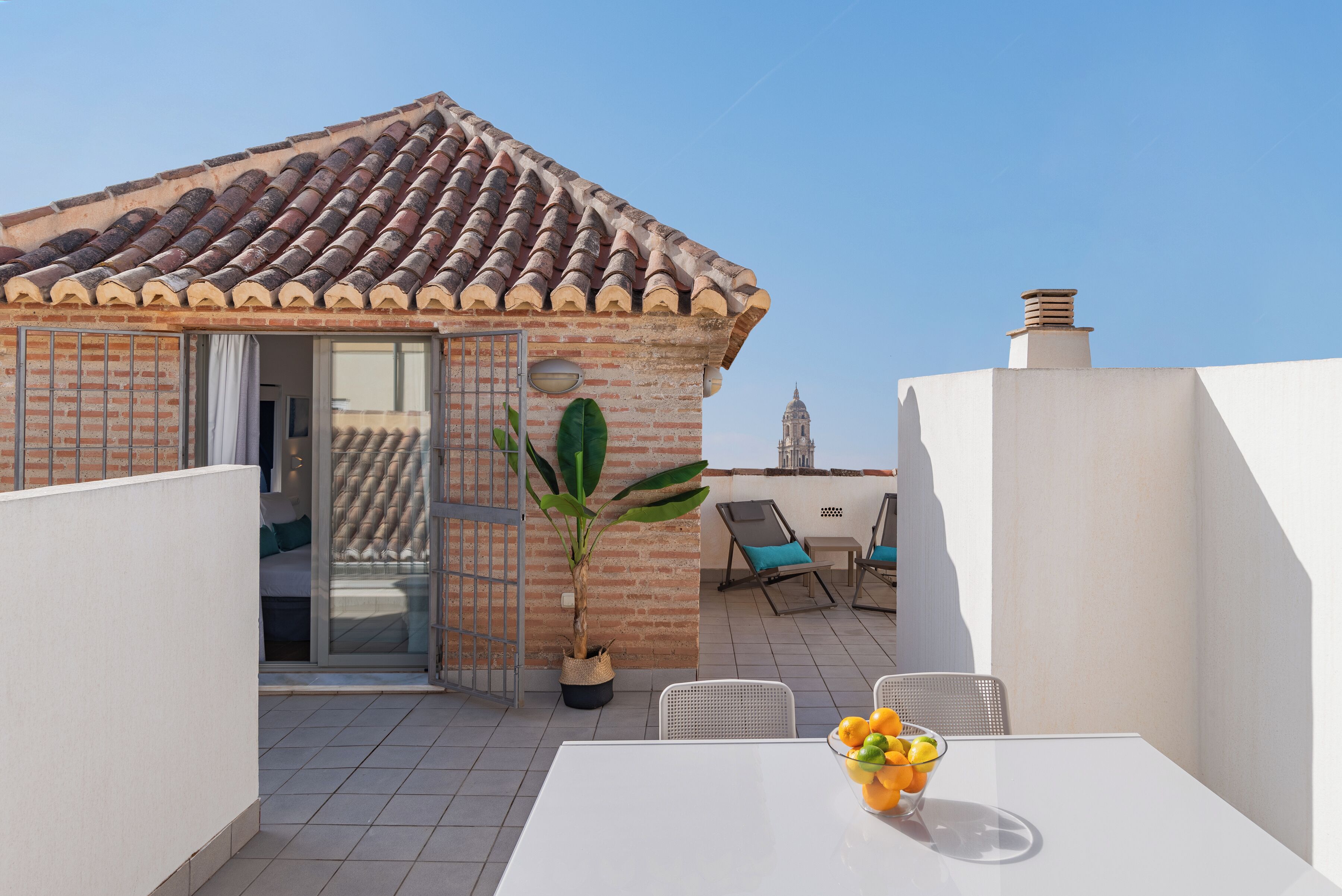 Eine Dachterrasse mit Ziegeldach, einem Blumentopf, einem Tisch mit Obstschale und im Hintergrund ein Blick auf ein Gebäude mit einem Glockenturm.