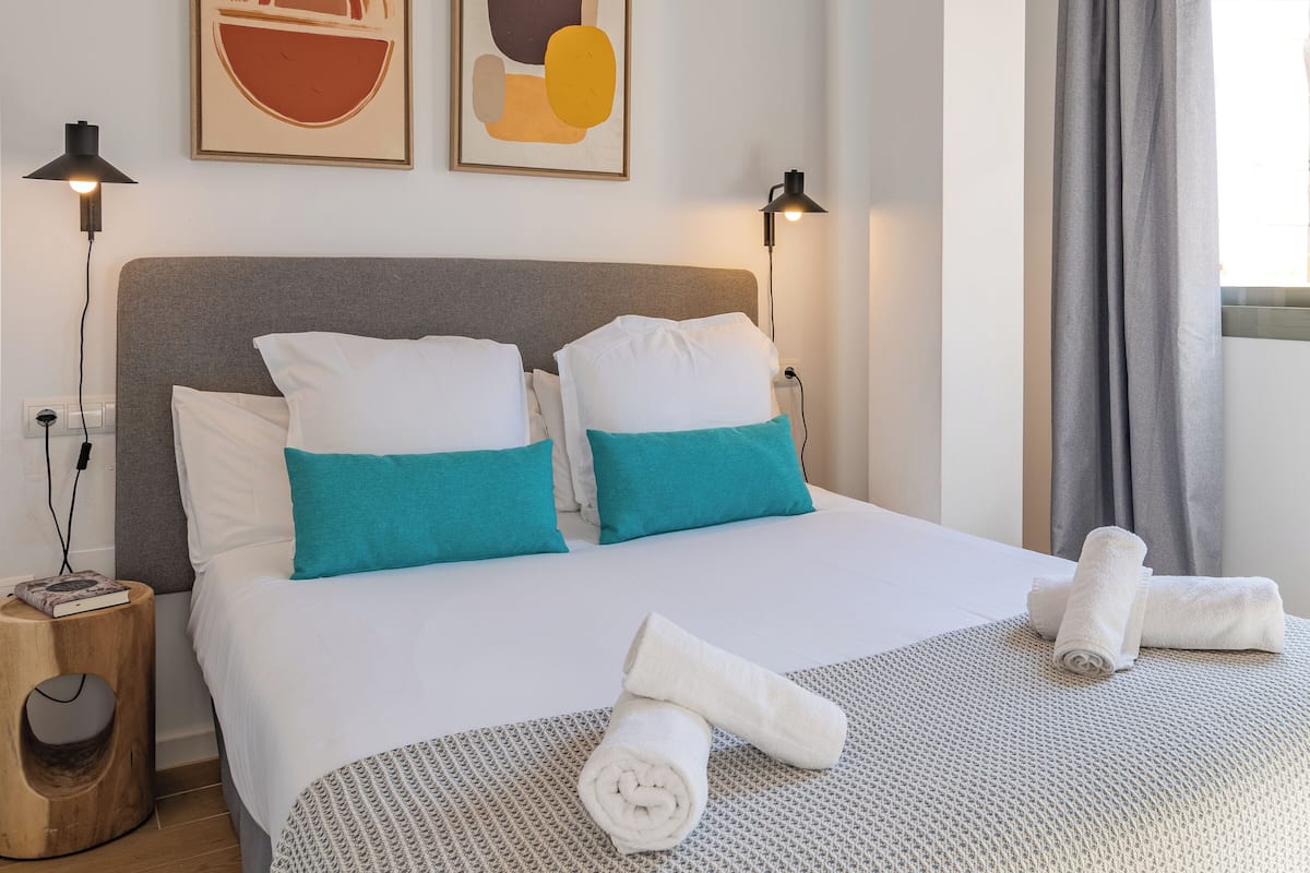 Una cama bien hecha con almohadas blancas y turquesa, una mesita de noche de madera con un libro y dos toallas enrolladas sobre una colcha con textura.