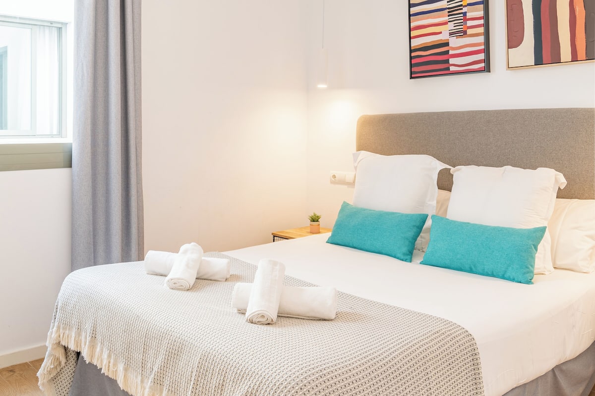 Una cama bien hecha con toallas enrolladas, un cabecero gris y dos almohadas color turquesa.