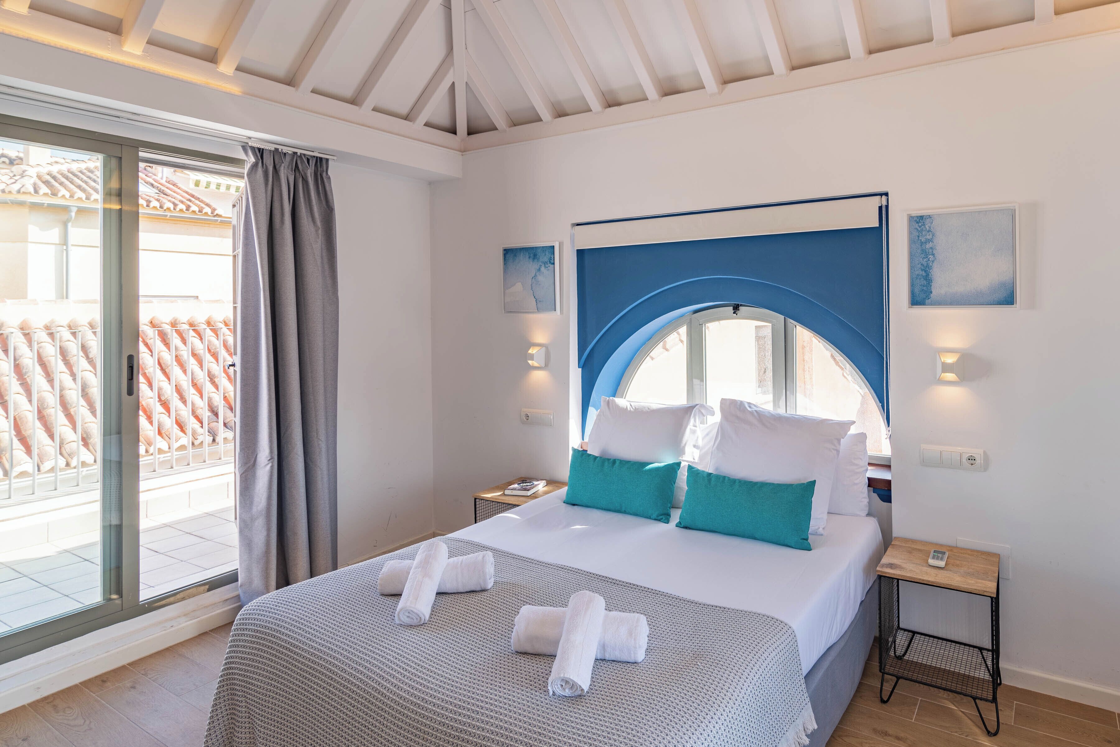 Une chambre d’hôtel avec un lit, une table de chevet, une fenêtre avec des rideaux et une vue sur un toit recouvert de tuiles rouges.
