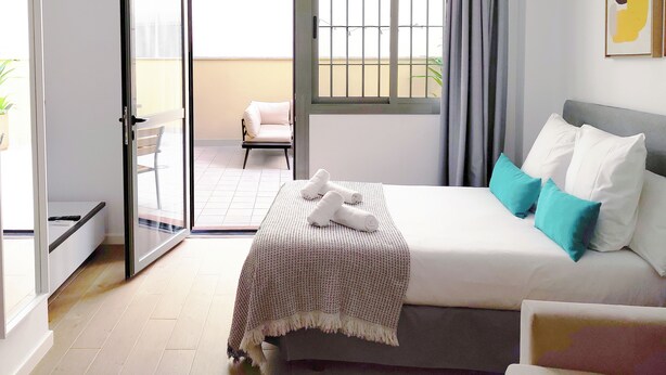A bedroom with a bed, a window with curtains, a balcony with a chair, and a door leading outside.