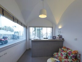 A modern living room with a large window, a sofa, a small table, and a kitchen area with a refrigerator and a clock.