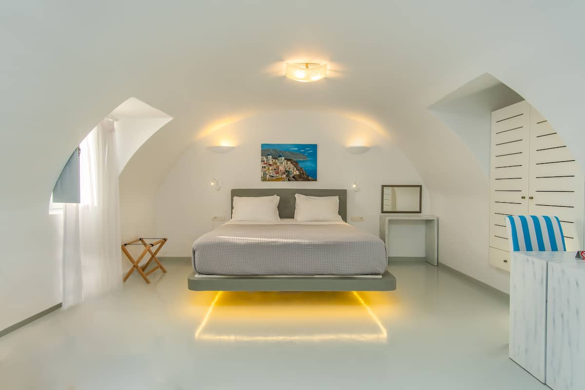 A modern bedroom with a bed centered under a sloped ceiling, a painting above the bed, and a small table with a chair to the right.