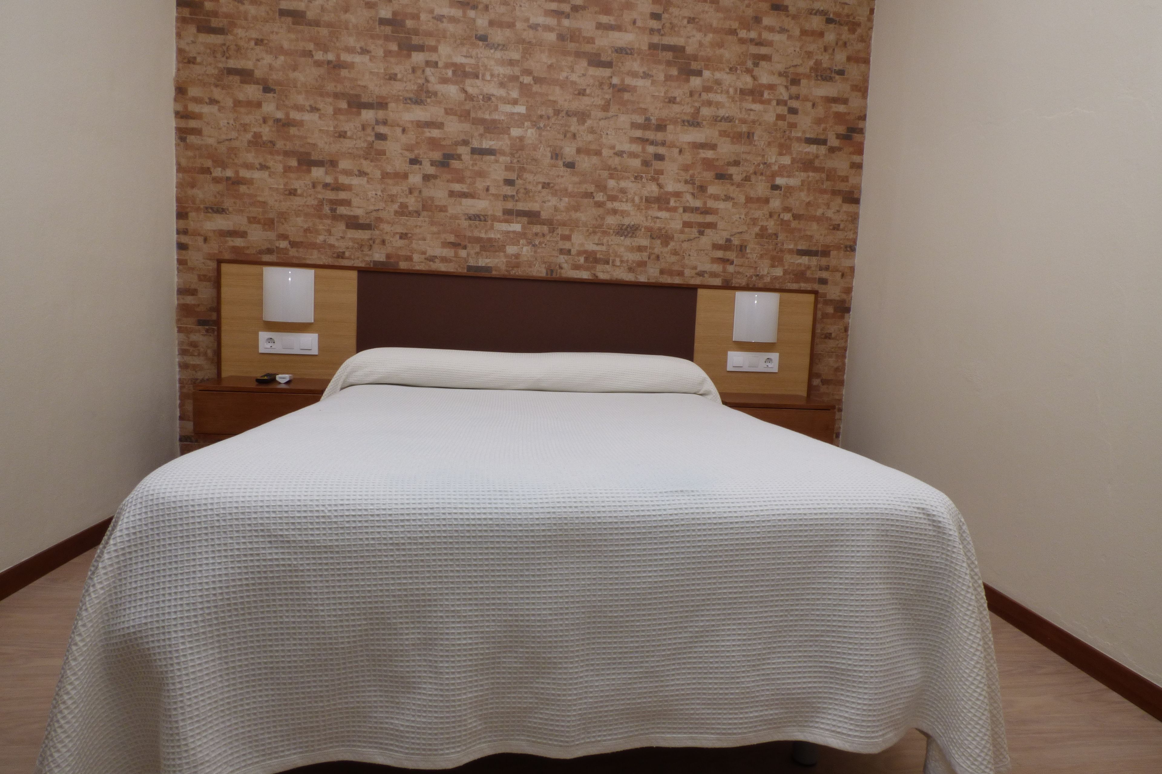 A neatly made bed with a white bedspread in a room with a brick-patterned wall and wooden headboard.