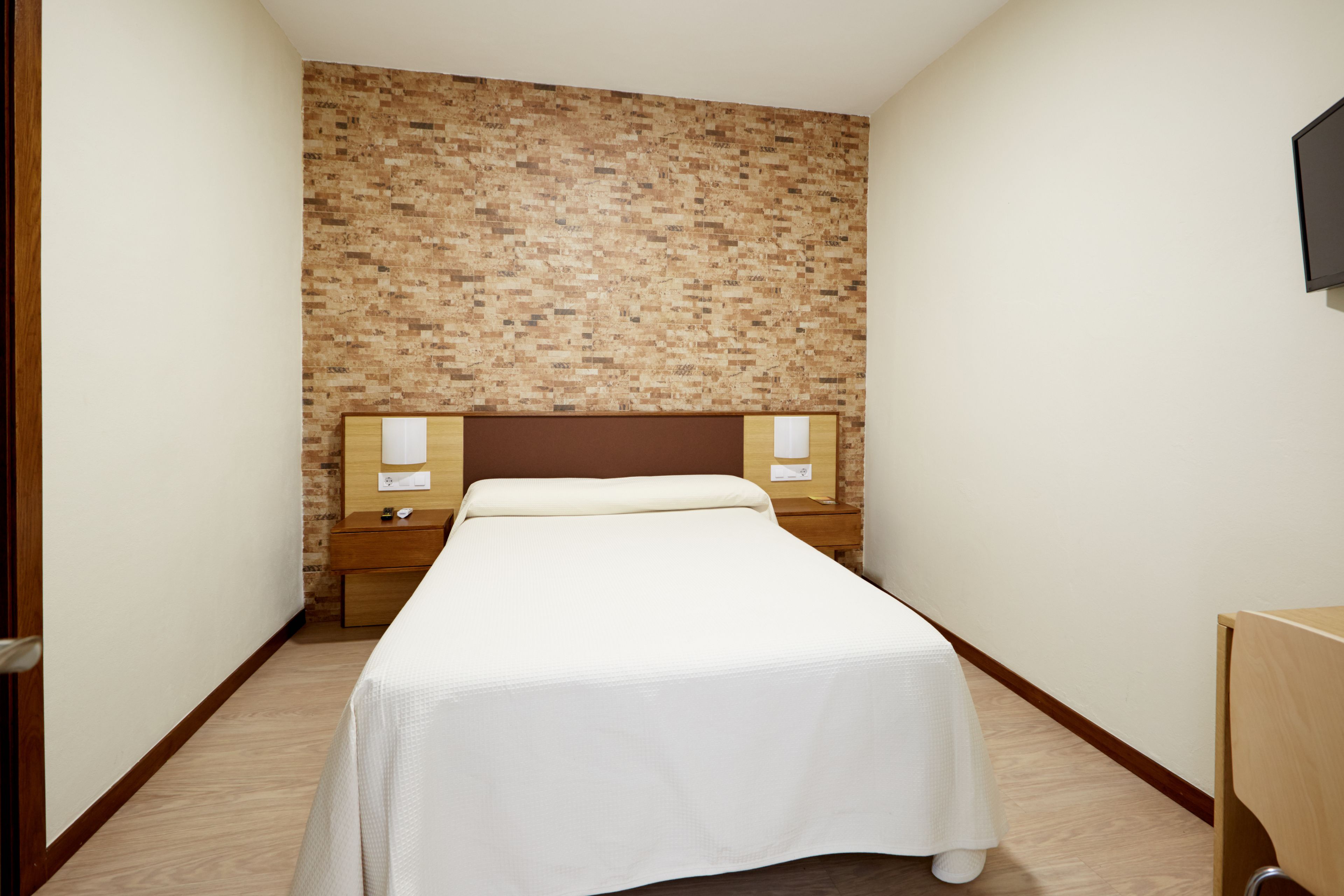 A hotel room with a brick-patterned wall, a wooden headboard, a white bedspread, and a nightstand with a lamp.