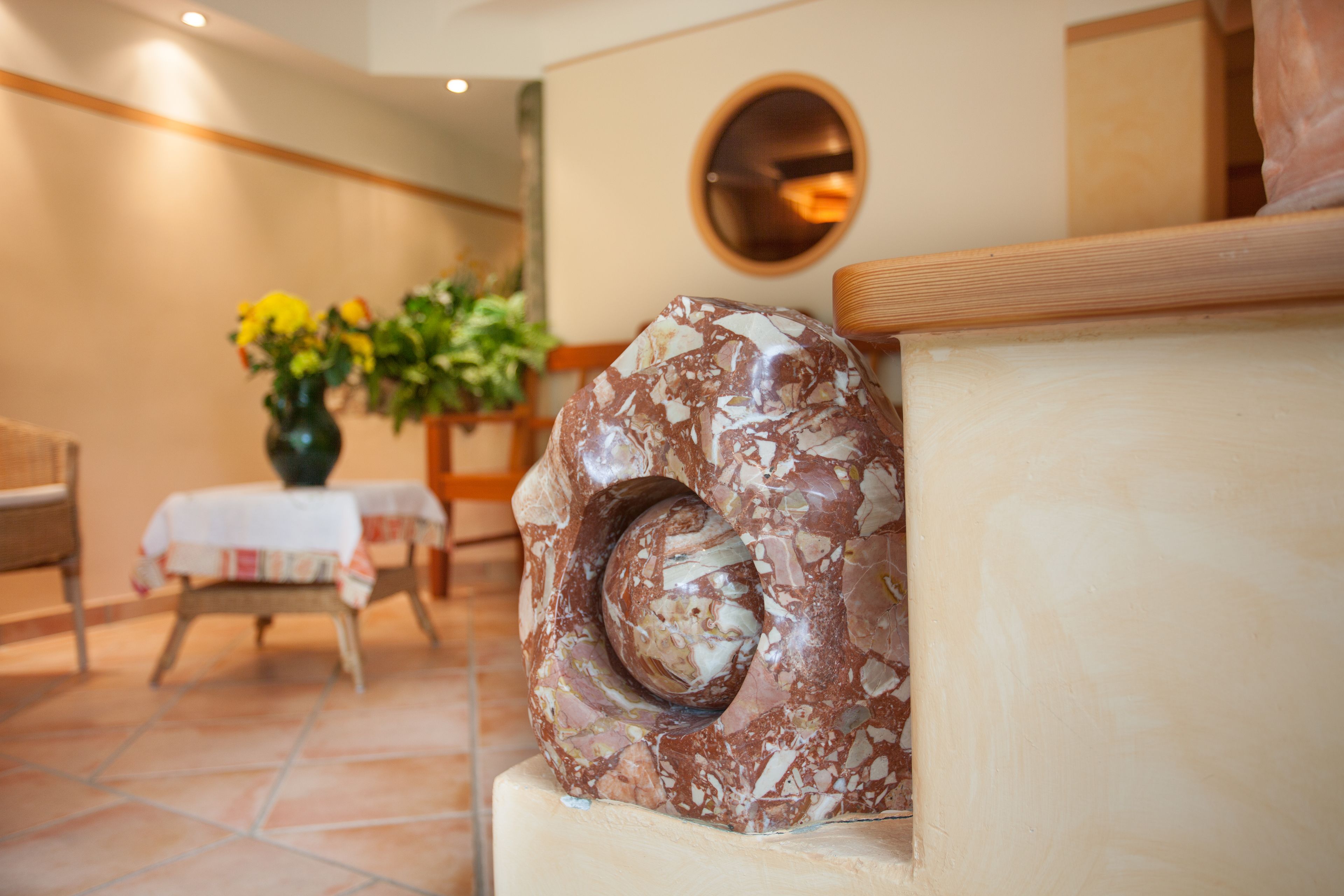 Une sculpture en marbre dans un hall d’entrée, avec un grand miroir rond au mur, une table ornée d’un vase de fleurs et un banc en bois.