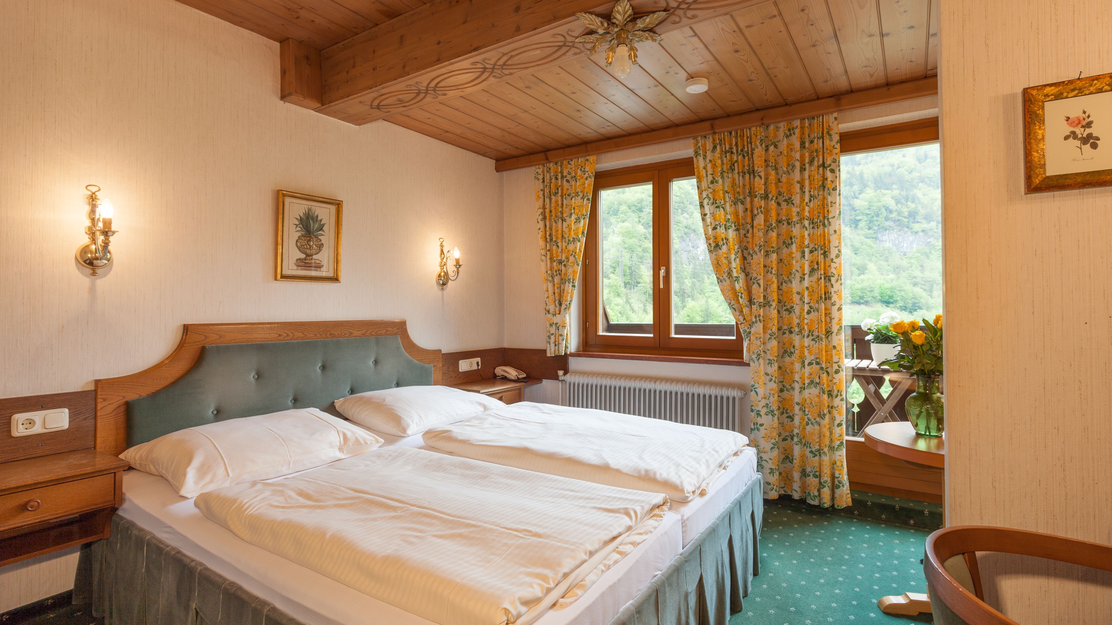 A hotel room with a large bed, a wooden headboard, a window with floral curtains, a view of greenery, and a small balcony with flowers.