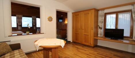 Une pièce avec un plancher en bois, un canapé, une table recouverte d’une nappe, un téléviseur posé sur un meuble et des armoires en bois.