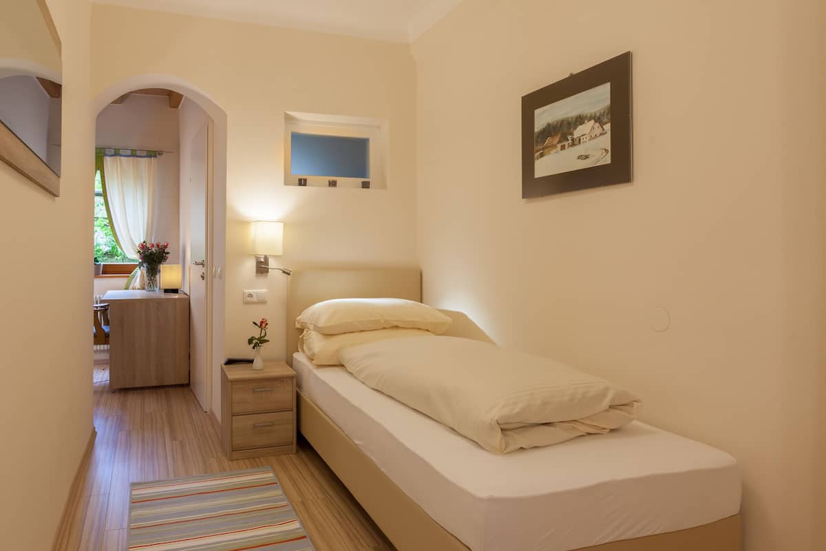 Een kleine, keurig opgemaakt slaapkamer met een enkel bed, nachtkastje en een nachtkast. Er is een raam met gordijnen, een klein schilderij aan de muur en een deur die naar een andere kamer leidt.