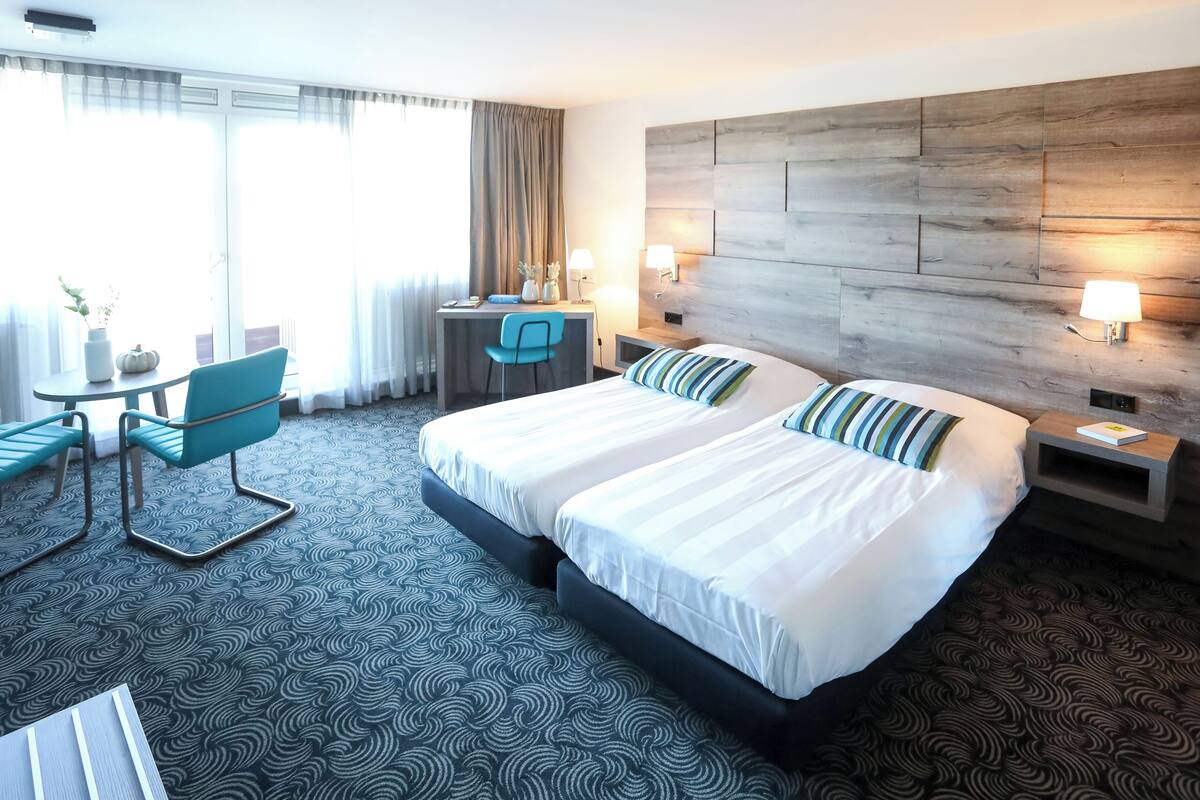 Een hotelkamer met een groot bed, een bureau met stoel, een klein tafeltje en een raam met gordijnen.