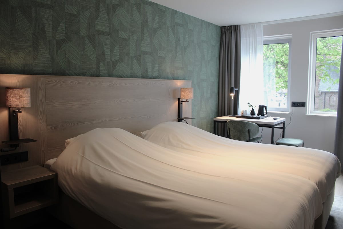 Een hotelkamer met een groot bed, twee bedlampjes, een bureau met stoel en een raam met uitzicht op groen.