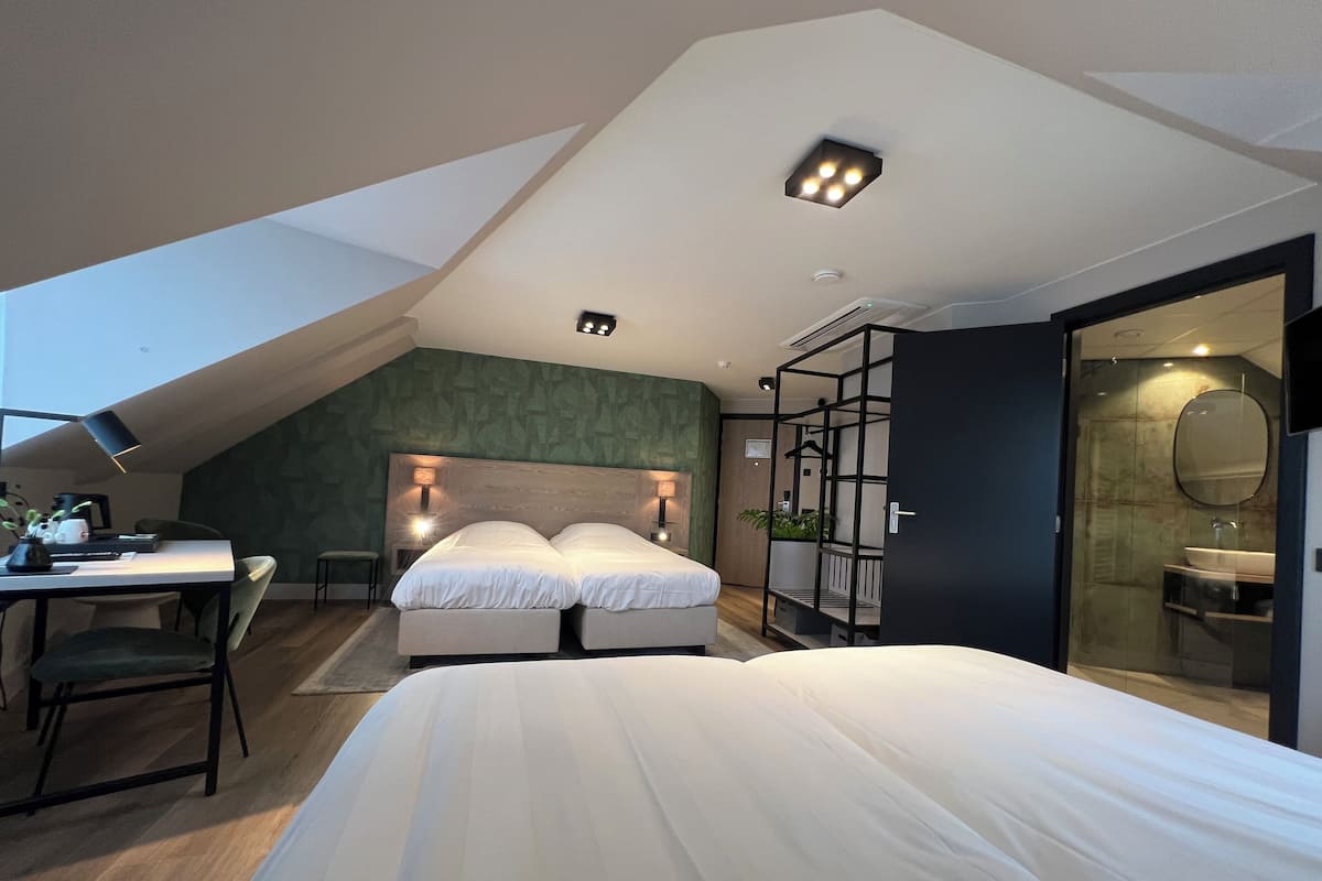 Een moderne slaapkamer met een schuine kap, een bed, een bureau met stoel, en een badkamer met spiegel en wastafel.