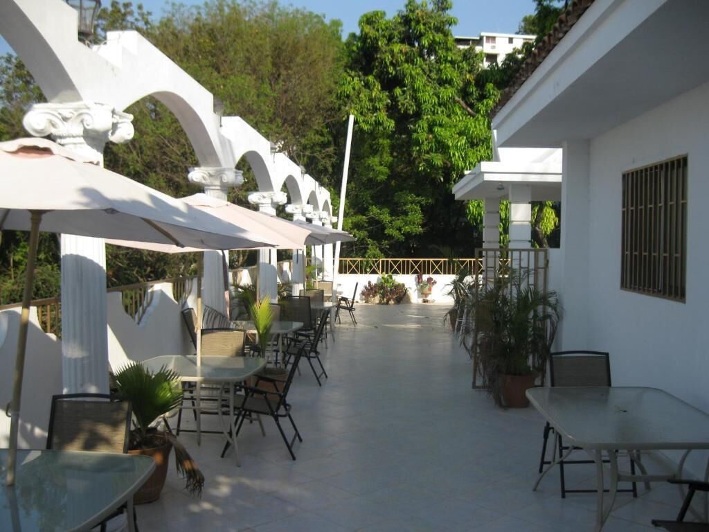 Une terrasse extérieure couverte, aménagée avec des tables et des chaises, entourée de murs blancs et de plantes en pot.
