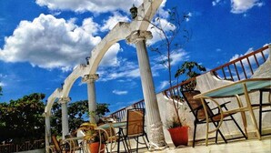 Área externa com assentos, colunas brancas e cadeiras sob um céu azul claro.