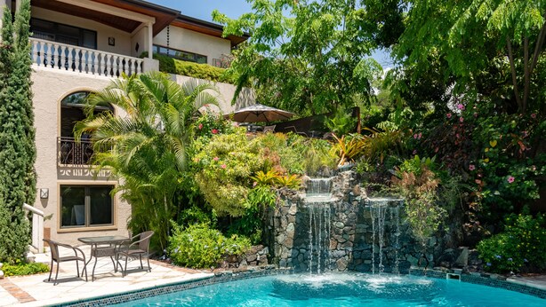 Una piscina con cascada, rodeada de abundante vegetación y un patio con mobiliario exterior.