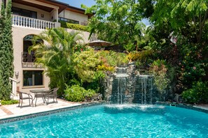 Una piscina con cascada, rodeada de abundante vegetación y un patio con mobiliario exterior.