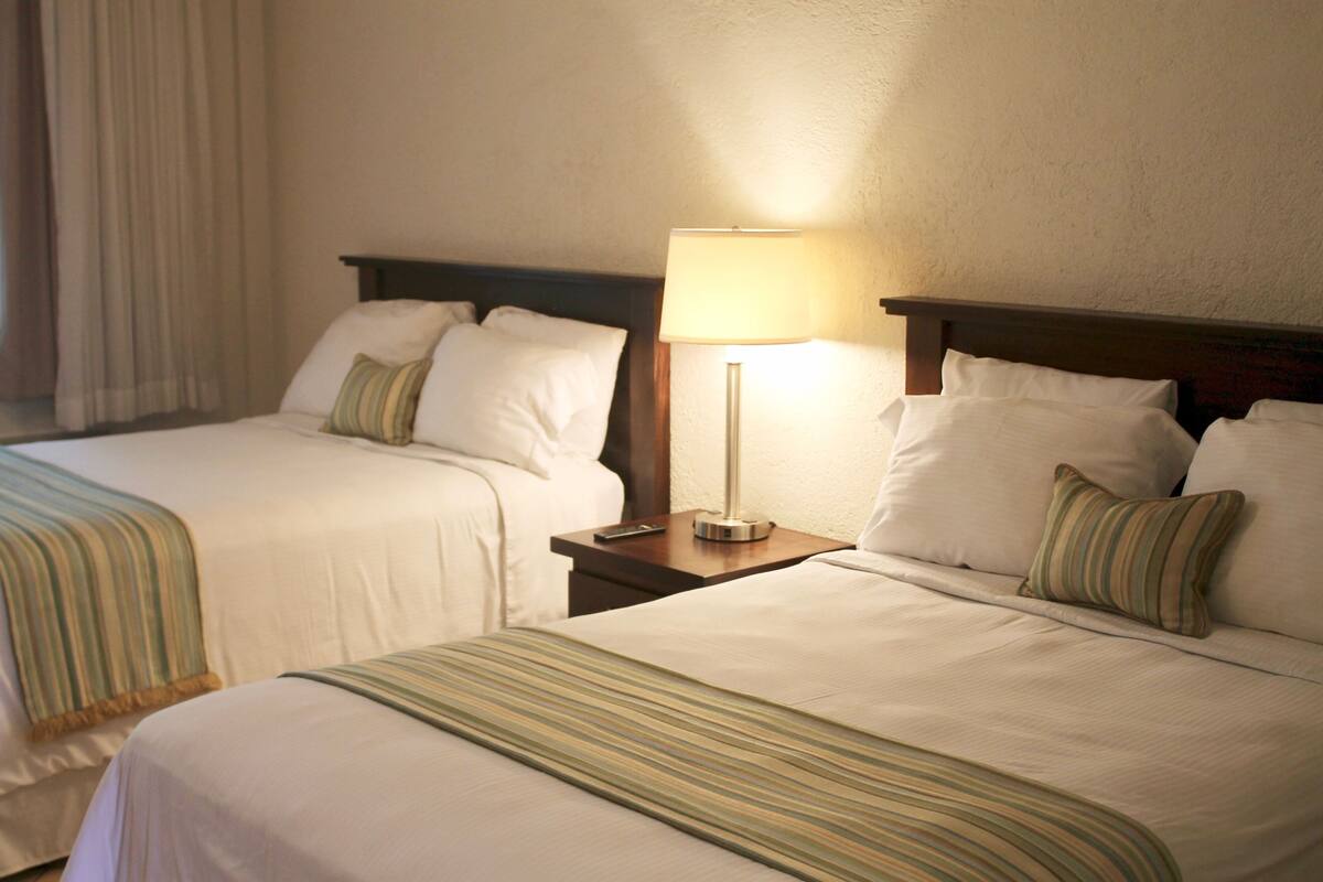 Two beds with white linens and striped blankets, a bedside table with a lamp, and a window with curtains.