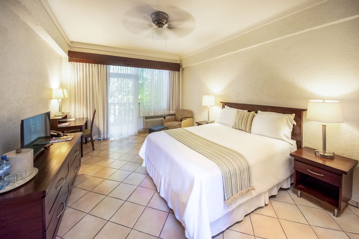 A hotel room with a large bed, two bedside tables with lamps, a desk with a chair, a television, and a window with curtains.