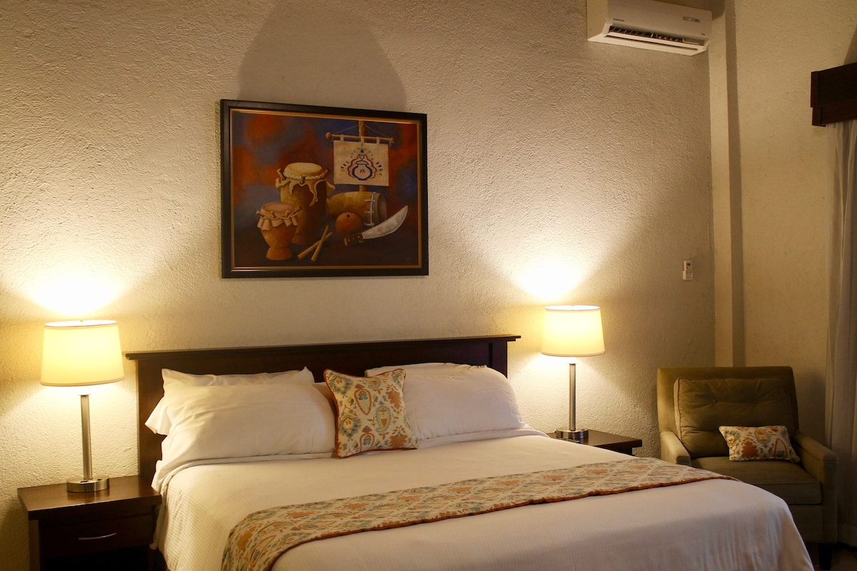 A neatly made bed with white linens, two bedside lamps, a chair with a cushion, and a wall-mounted air conditioner.