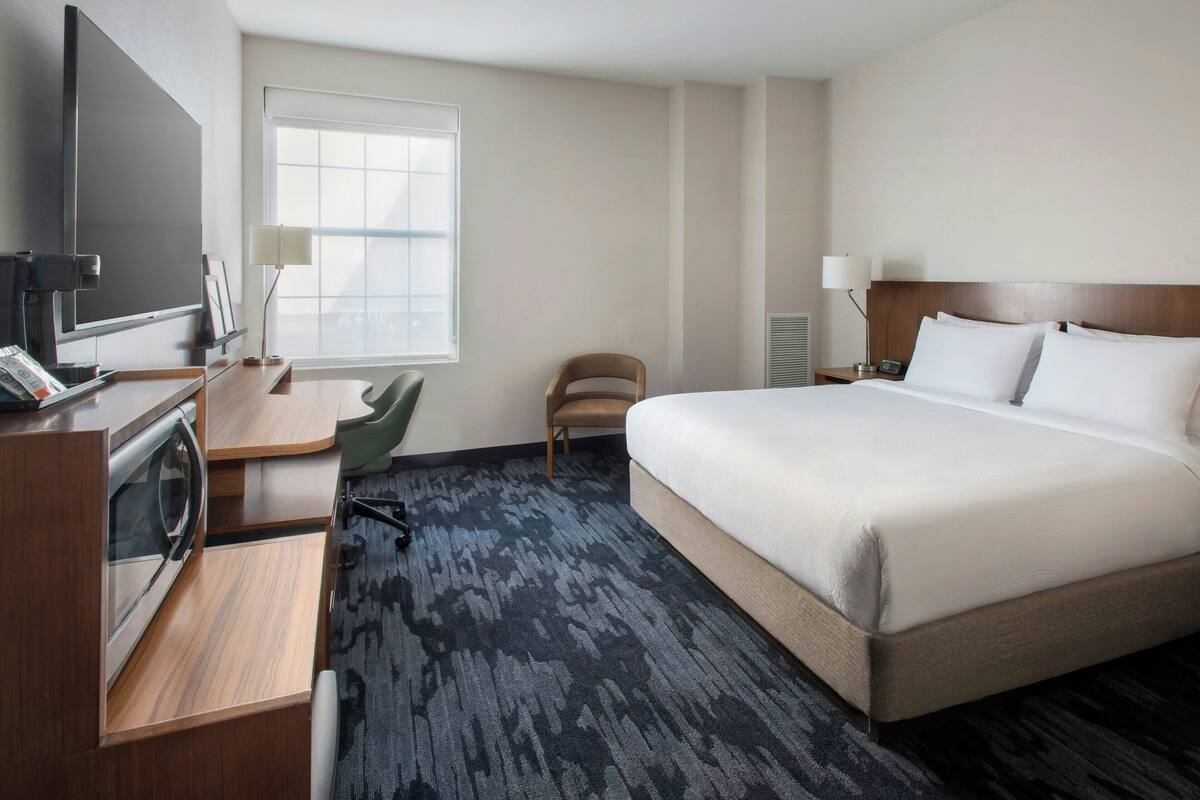 A hotel room with a bed, a desk with a computer, a chair, a TV, and a window.