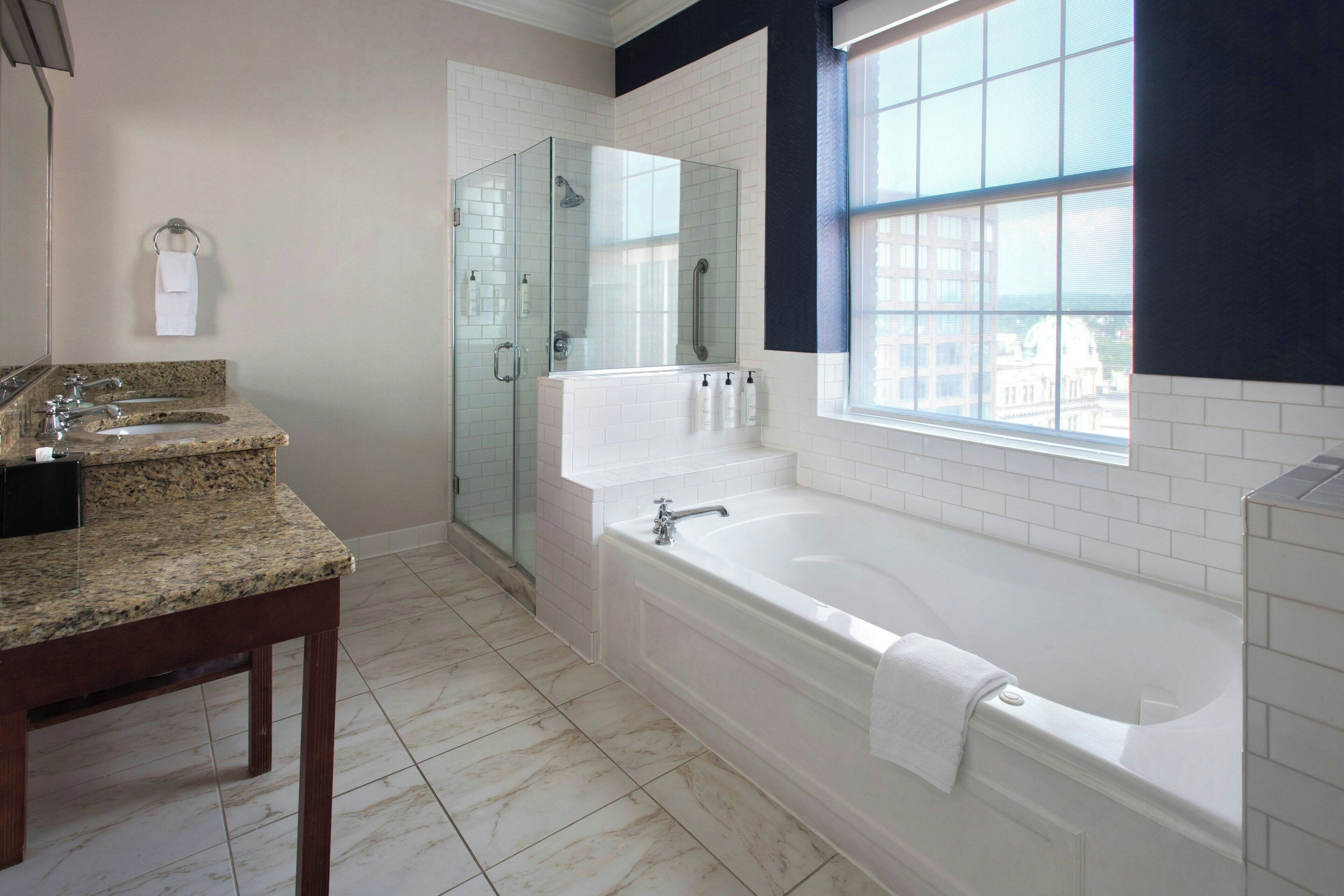 A modern bathroom with a granite countertop, a freestanding bathtub, a glass-enclosed shower, and a window providing a view of the city.