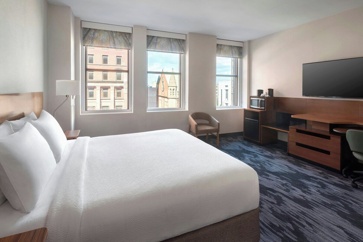 Ein Hotelzimmer mit einem großen Bett, einem Schreibtisch, einem Flachbildfernseher, einem Sessel und Blick auf Gebäude durch die Fenster.