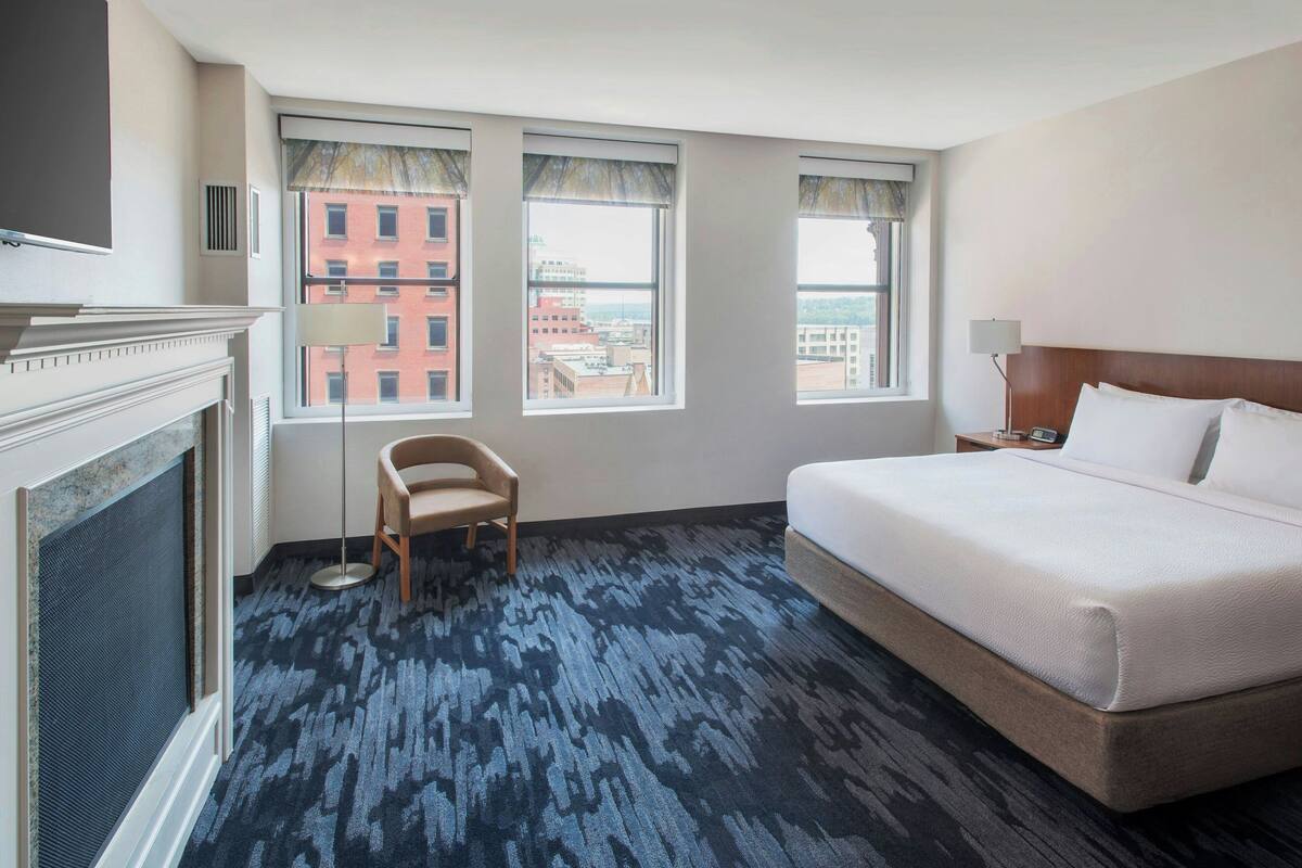 Ein Hotelzimmer mit einem großen Bett, einem Sessel, einem Kamin und drei Fenstern mit Blick auf die Stadt.