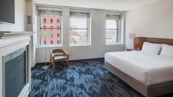 A hotel room with a large bed, a chair, a fireplace, and three windows with city views.