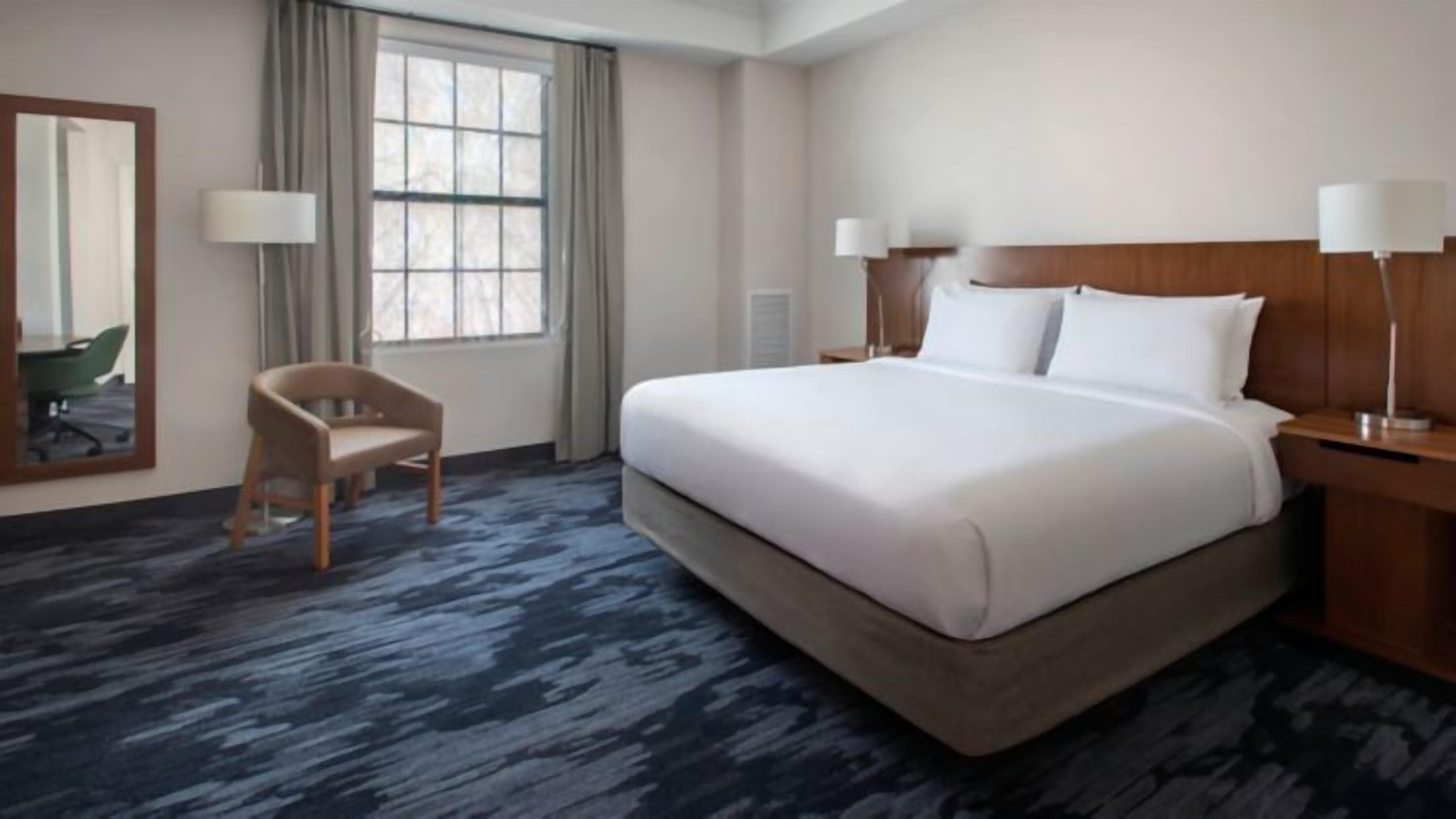A hotel room with a large bed, a chair, a desk, a lamp, and a window.